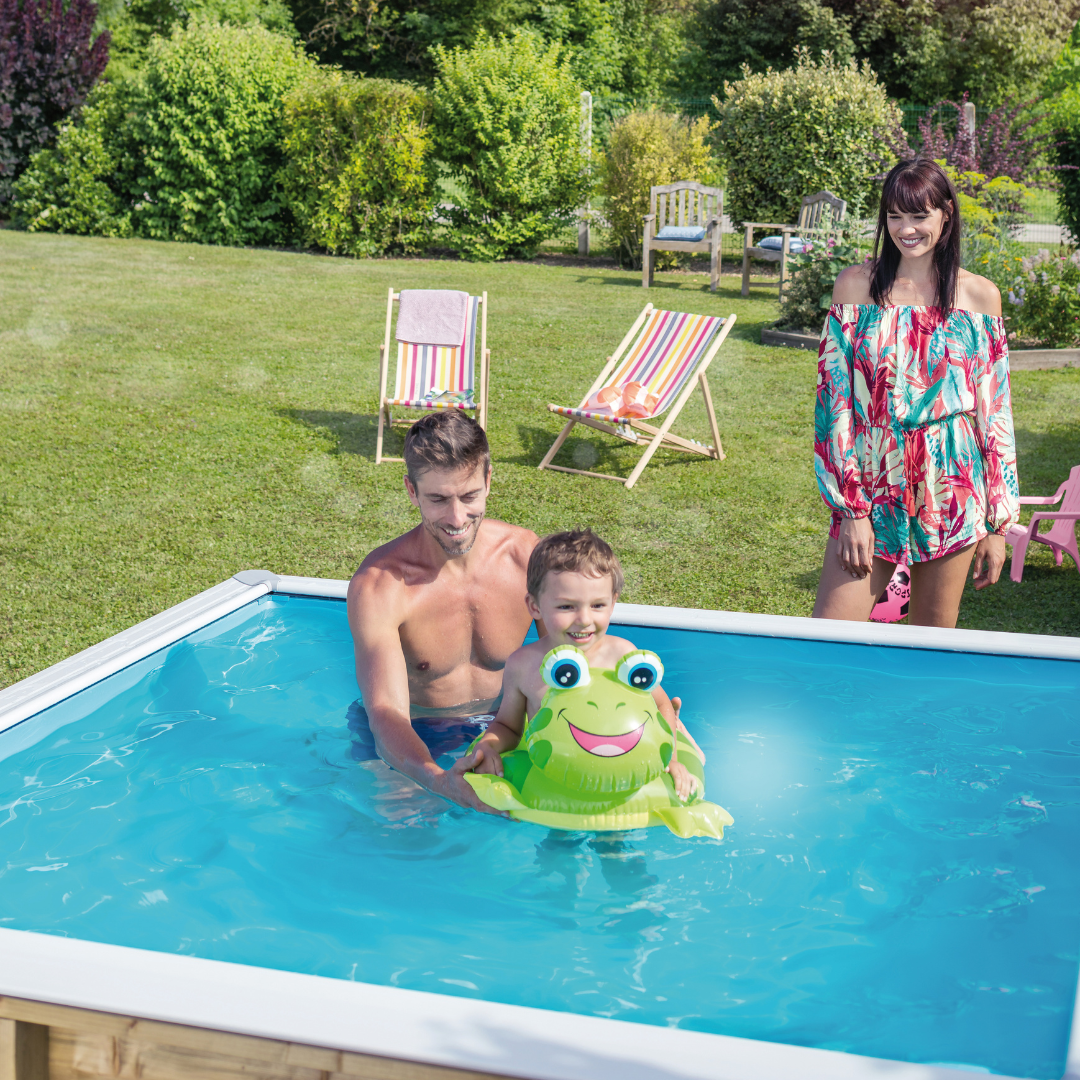 Familia en una piscina de madera Pistoche al aire libre, padre y niño con un flotador en forma de rana, mientras la madre observa sonriente.
