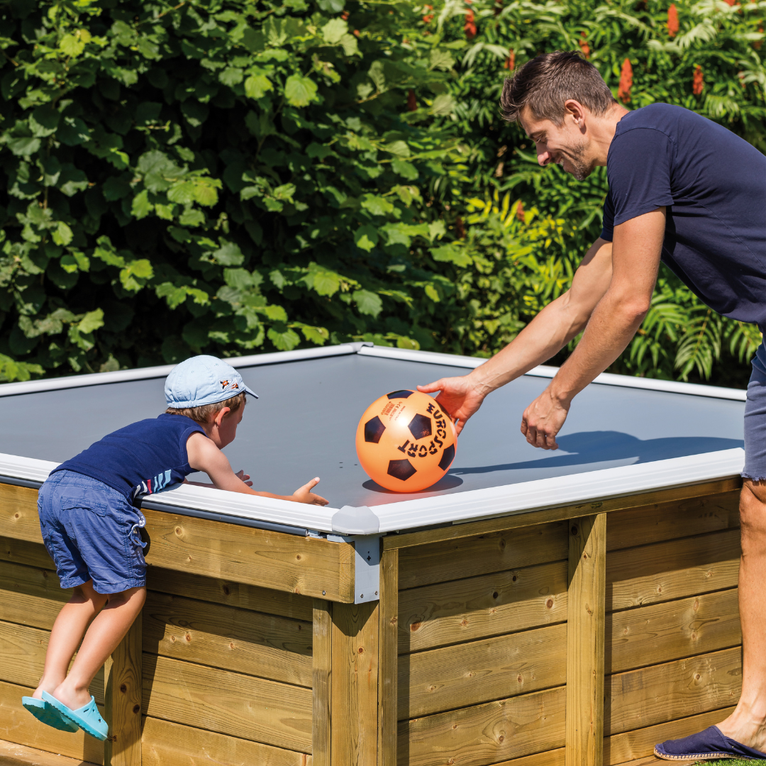 Padre e hijo jugando un juego de balón en una piscina de madera Pistoche, con el niño apoyado en el borde y el padre sonriendo mientras golpea una pelota naranja.