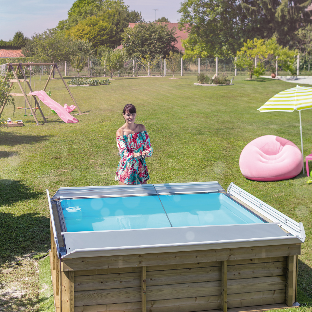 Piscina de madera Pistoche en jardín, mujer con bikini coloreado junto a la piscina desmontable rodeada de césped y juegos infantiles.