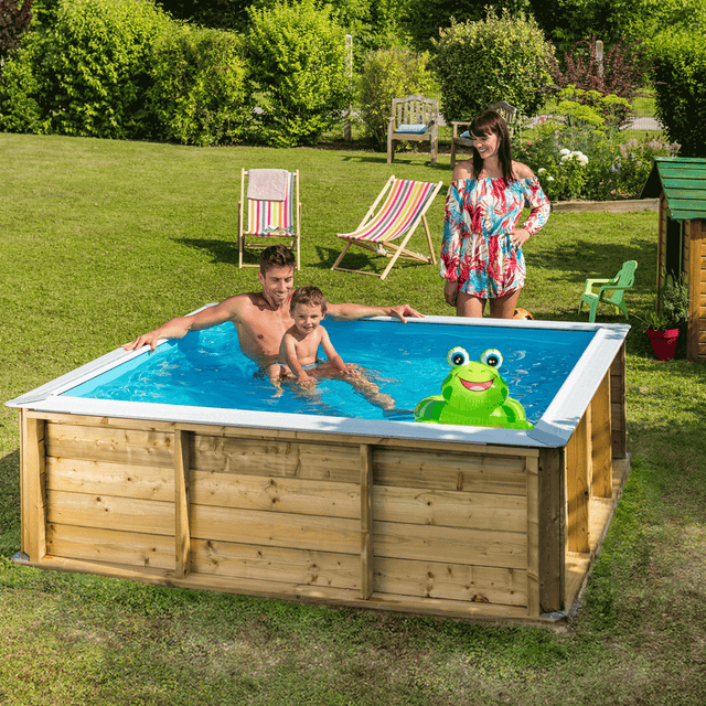 Piscina de madera Pistoche en jardín, familia jugando en el agua con juguete de rana inflable junto a sillas y casa de juegos.